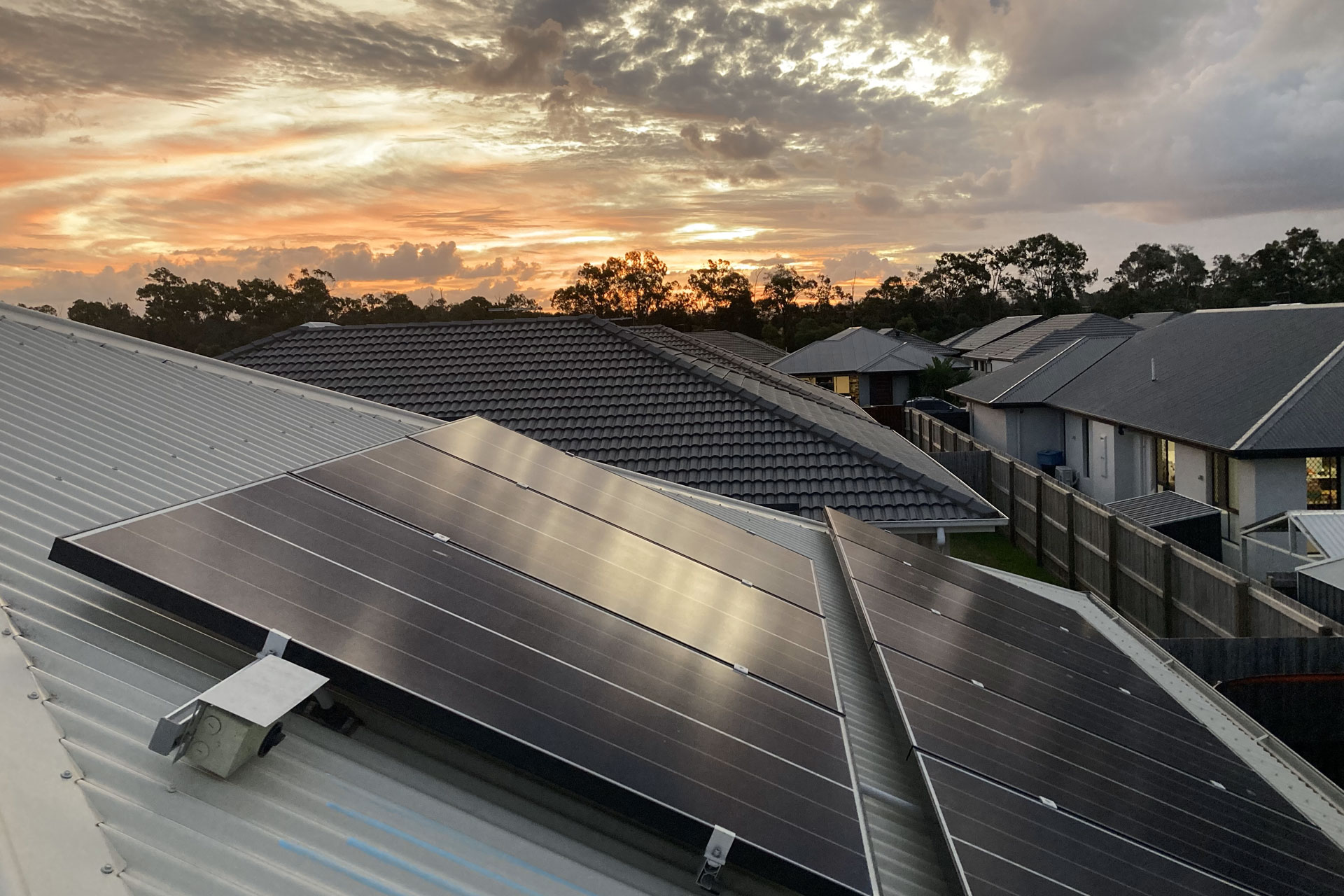 Rooftop solar panels, installed by Cairns solar specialists, gleam on a metal roof at sunset, with neighboring homes and lush tropical trees beneath a vibrant, cloud-streaked sky.