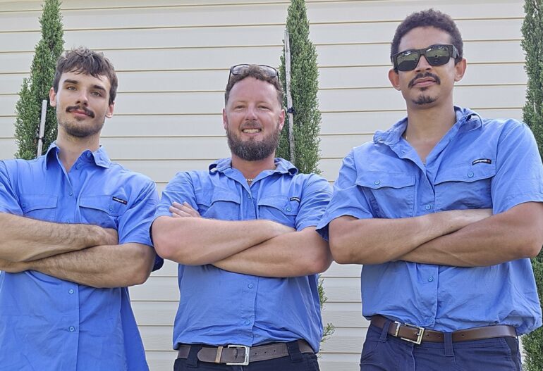 Three men in blue work shirts and shorts stand smiling with arms crossed in front of a light-colored building and tall, narrow bushes—ready to install commercial solar power systems Cairns and other Far North Queensland suburbs can depend on.