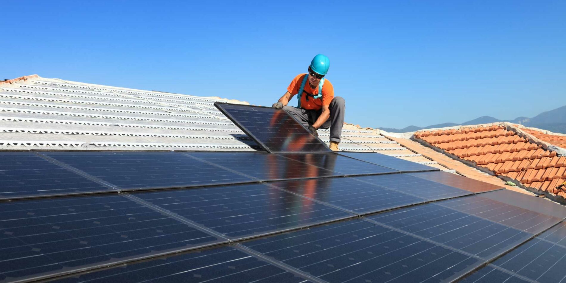 In Cairns, a worker wearing a blue helmet and orange vest uses SolaProtect Rooftop Assist to install solar panels on a tiled roof beneath a clear blue tropical sky, with the lush mountains of Far North Queensland visible in the background.