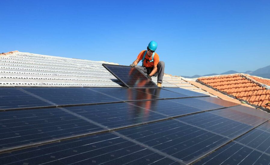 In Cairns, a worker wearing a blue helmet and orange vest uses SolaProtect Rooftop Assist to install solar panels on a tiled roof beneath a clear blue tropical sky, with the lush mountains of Far North Queensland visible in the background.