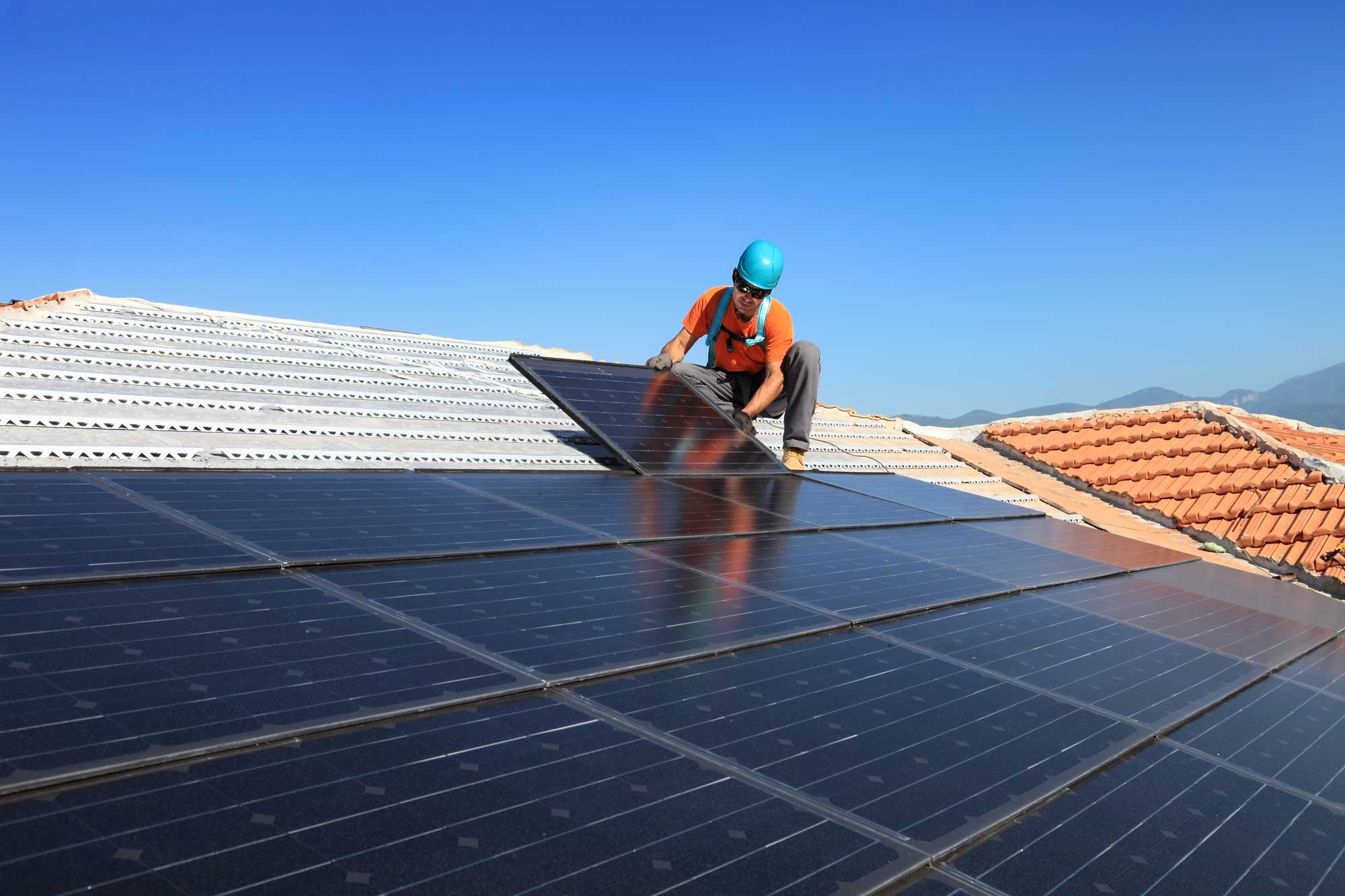 In Cairns, a worker wearing a blue helmet and orange vest uses SolaProtect Rooftop Assist to install solar panels on a tiled roof beneath a clear blue tropical sky, with the lush mountains of Far North Queensland visible in the background.