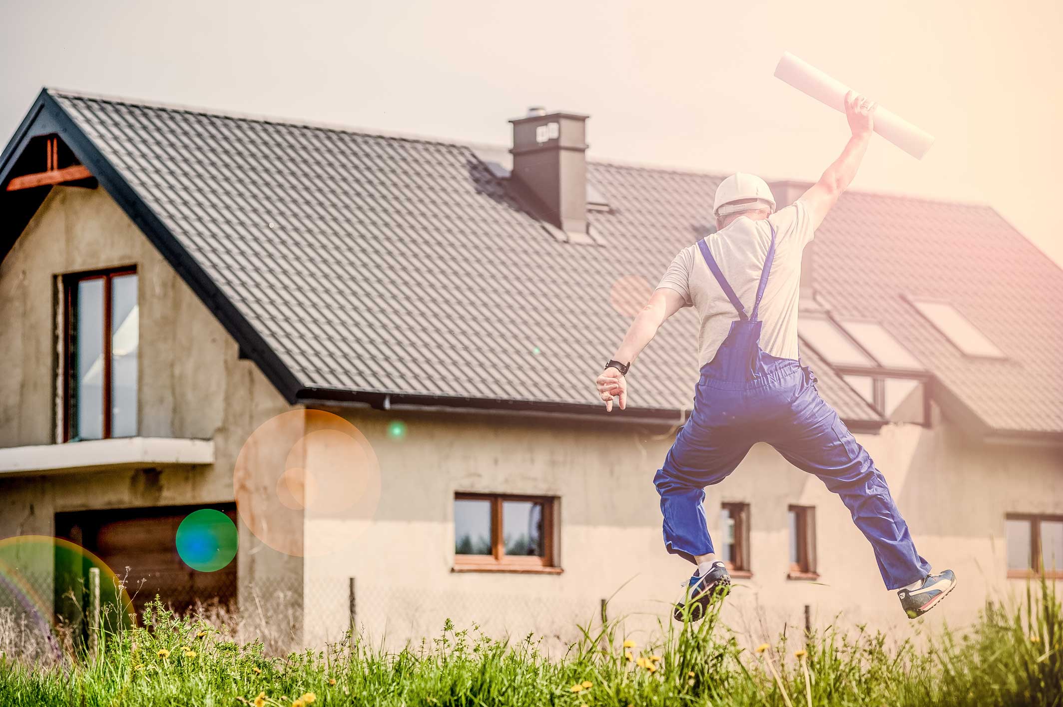A construction worker in blue overalls and a helmet jumps with joy while holding blueprints in front of a modern house in Cairns, Far North Queensland.