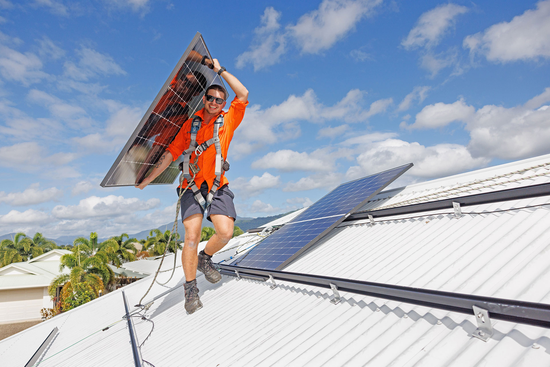 A smiling worker in orange safety gear carries a solar panel on a rooftop in Cairns, with palm trees and solar panels behind.