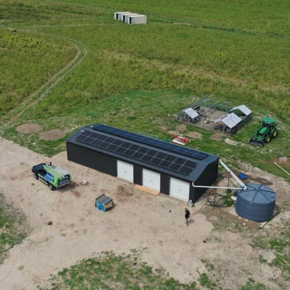 Aerial view of a black building with solar panels near Cairns, set in grassy fields. White car, green tractor, tanks, and farm gear nearby.