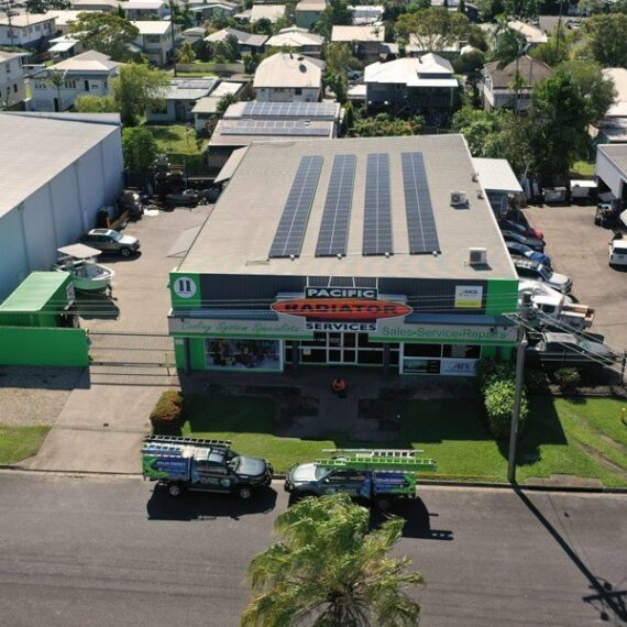 Aerial view of Pacific Radiators in Cairns with rooftop solar panels, two branded vehicles out front, and nearby FNQ buildings.