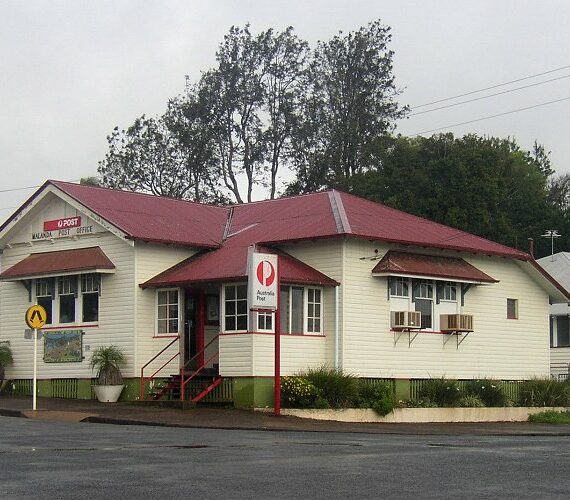 A small, white wooden post office with a red roof on a Cairns street corner, surrounded by tropical trees and road signs, under cloud cover.