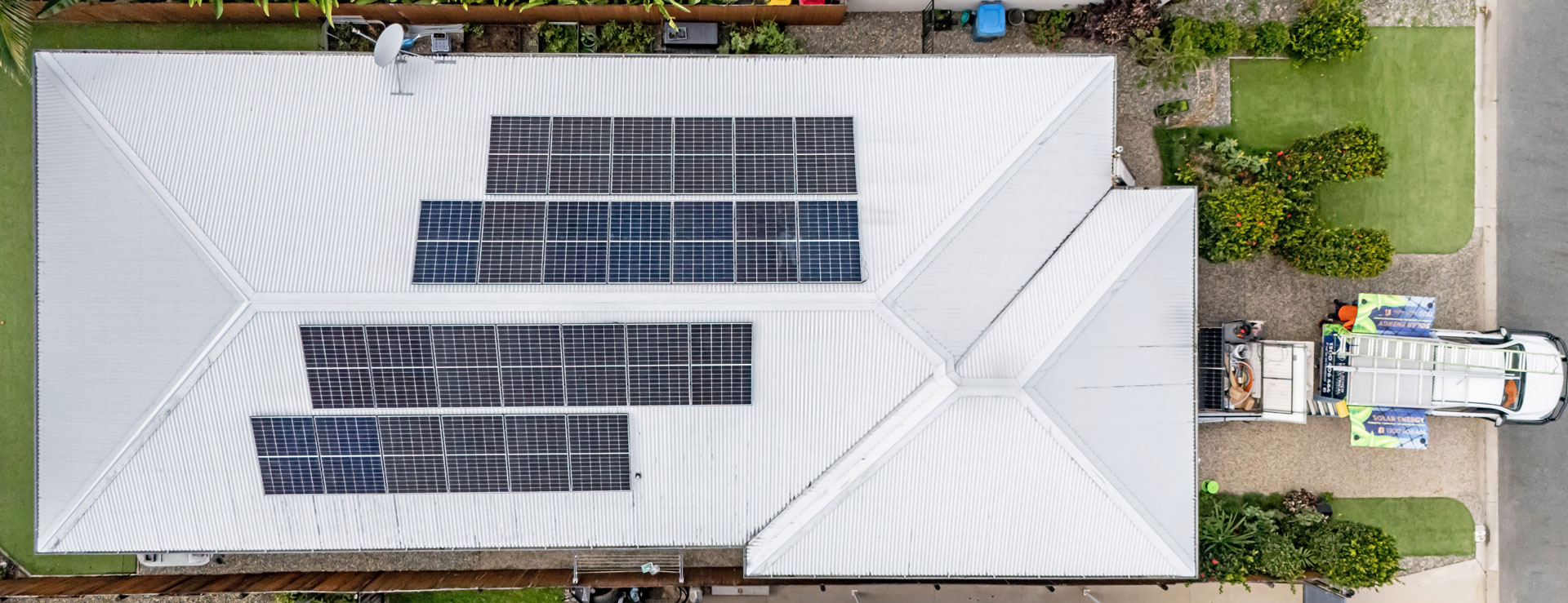 Aerial view of a white-roofed house with solar panels in Cairns, surrounded by lush green lawns and a driveway with a parked car.