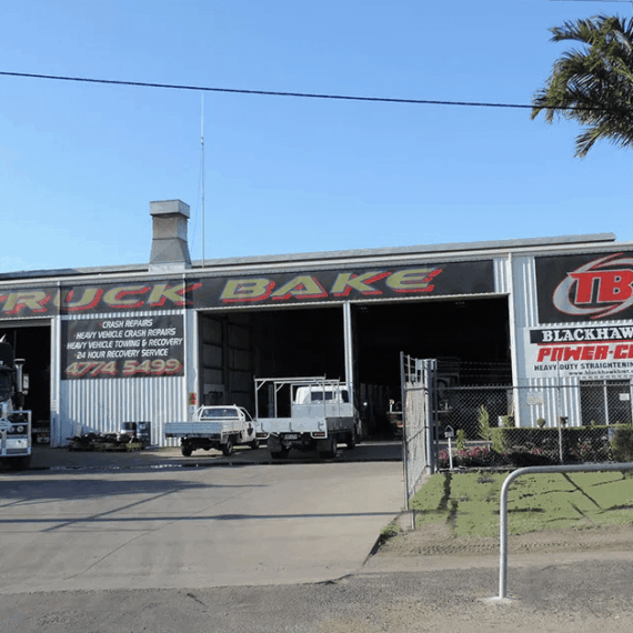A truck repair and painting workshop in Cairns with a "Truck Bake" sign, trucks parked around, palm trees, and chain-link fence.